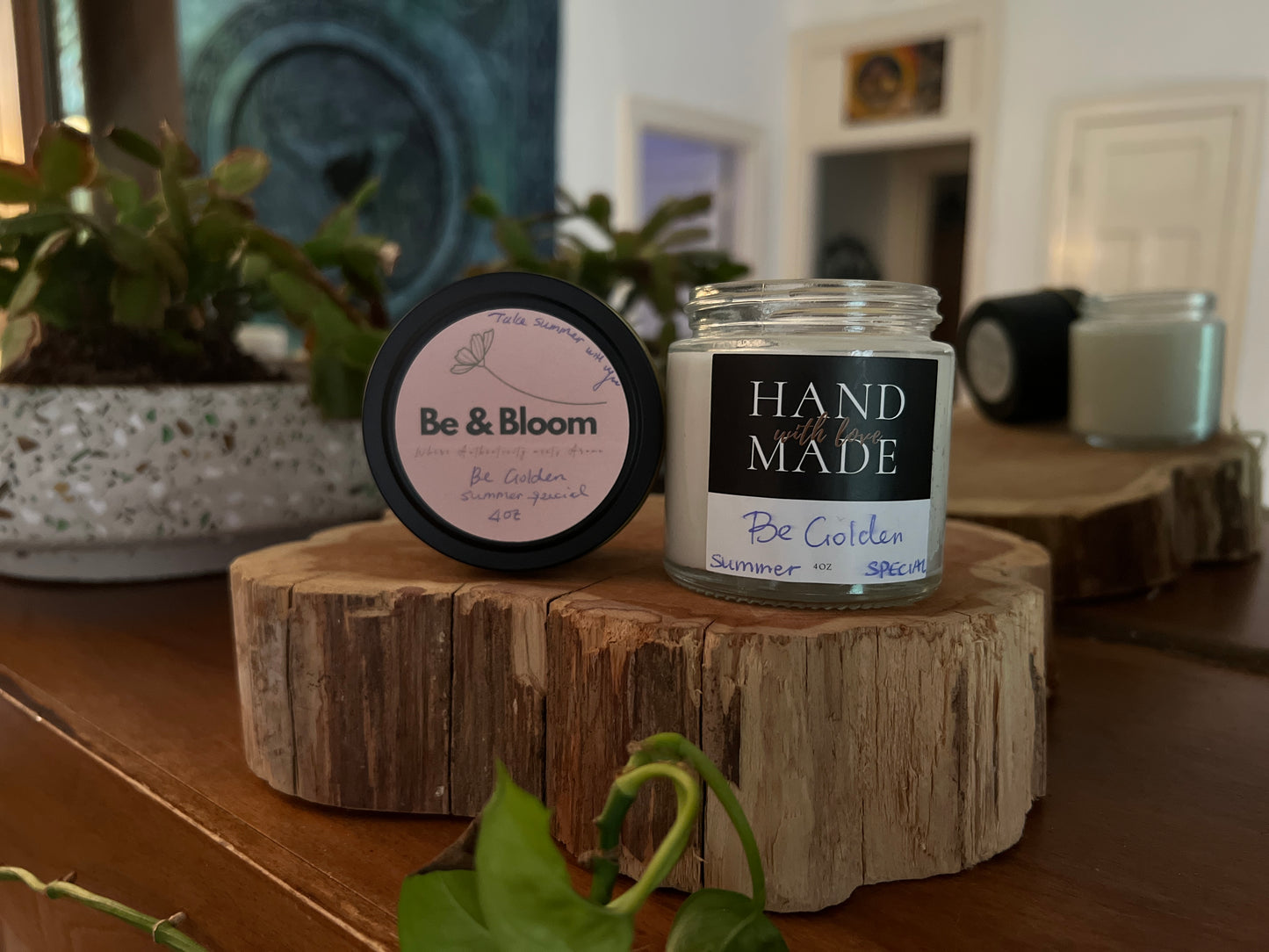 Jar and container of 'Be & Bloom' products on a wooden surface with plants in the background.